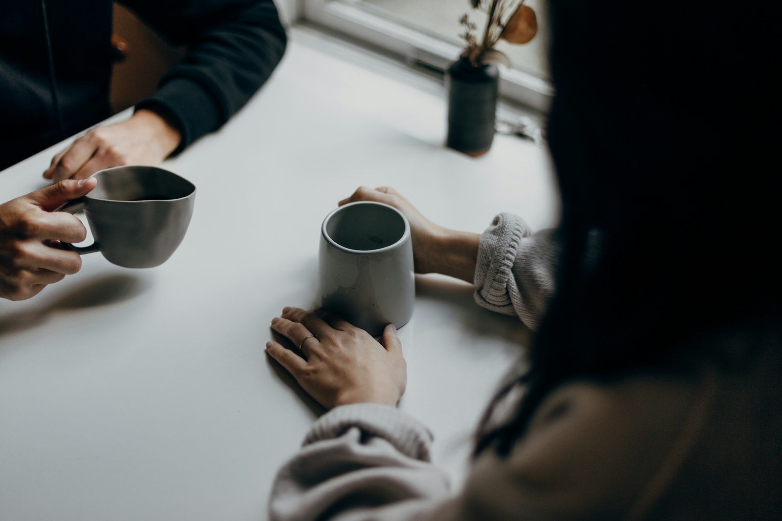 Two people holding gray mugs across a white table, mid-conversation